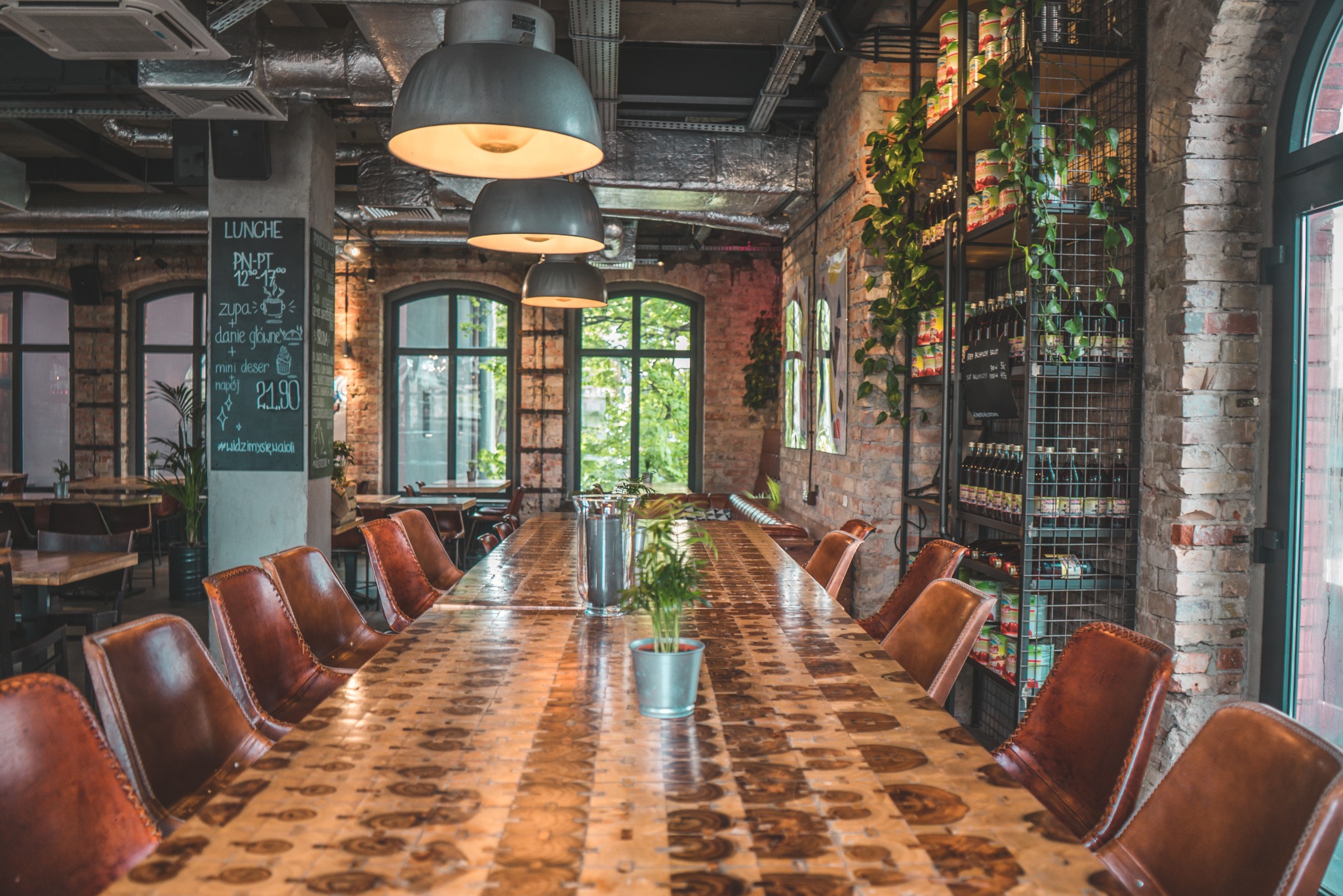 Wnętrze restauracji w stylu industrialnym z długim drewnianym stołem, skórzanymi krzesłami, ceglanymi ścianami, metalowymi regałami z napojami i roślinami pnącymi, oraz tablicą z menu lunchowym.