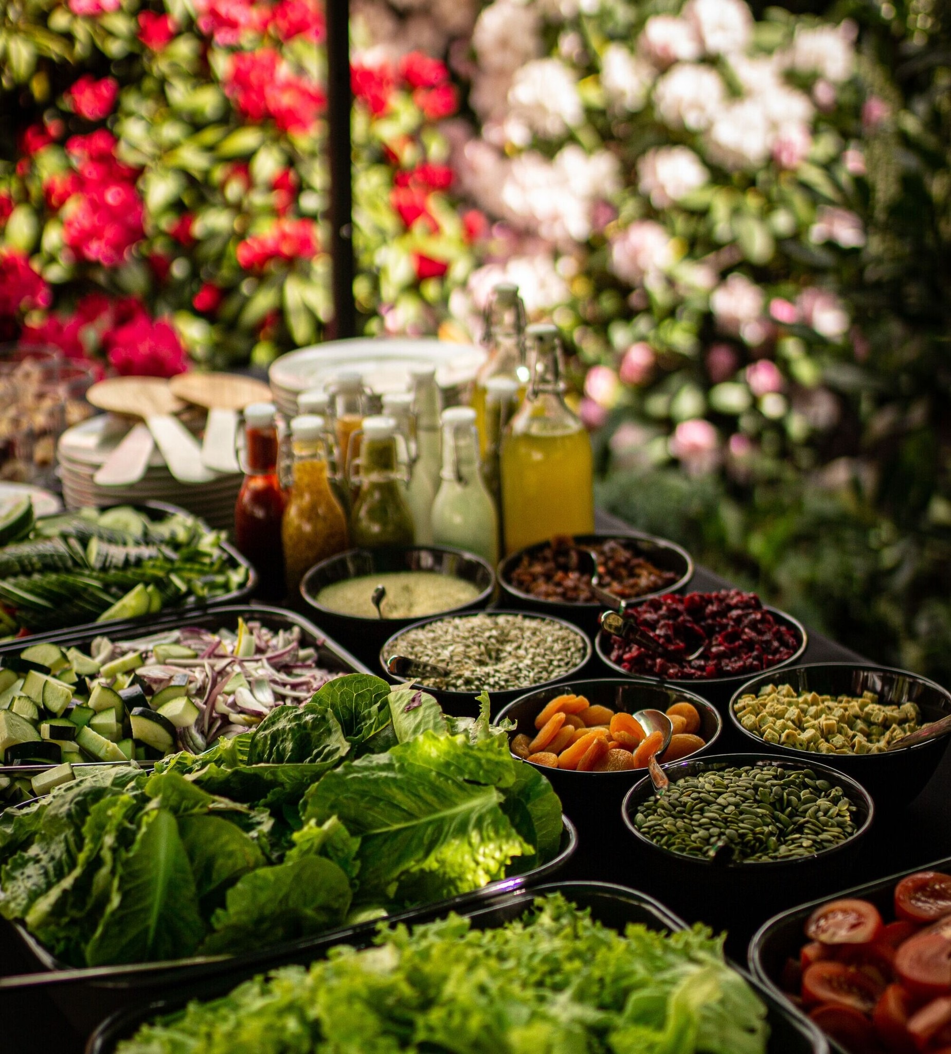 Stół z różnorodnymi składnikami sałatkowymi w czarnych misach i butelkach z sosami, ustawiony na tle kwitnących krzewów.