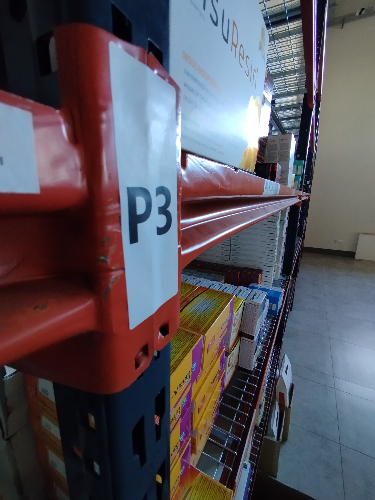 Półka regałowa w magazynie z oznaczeniem 'P3', wypełniona kartonami z lekami i suplementami diety.
