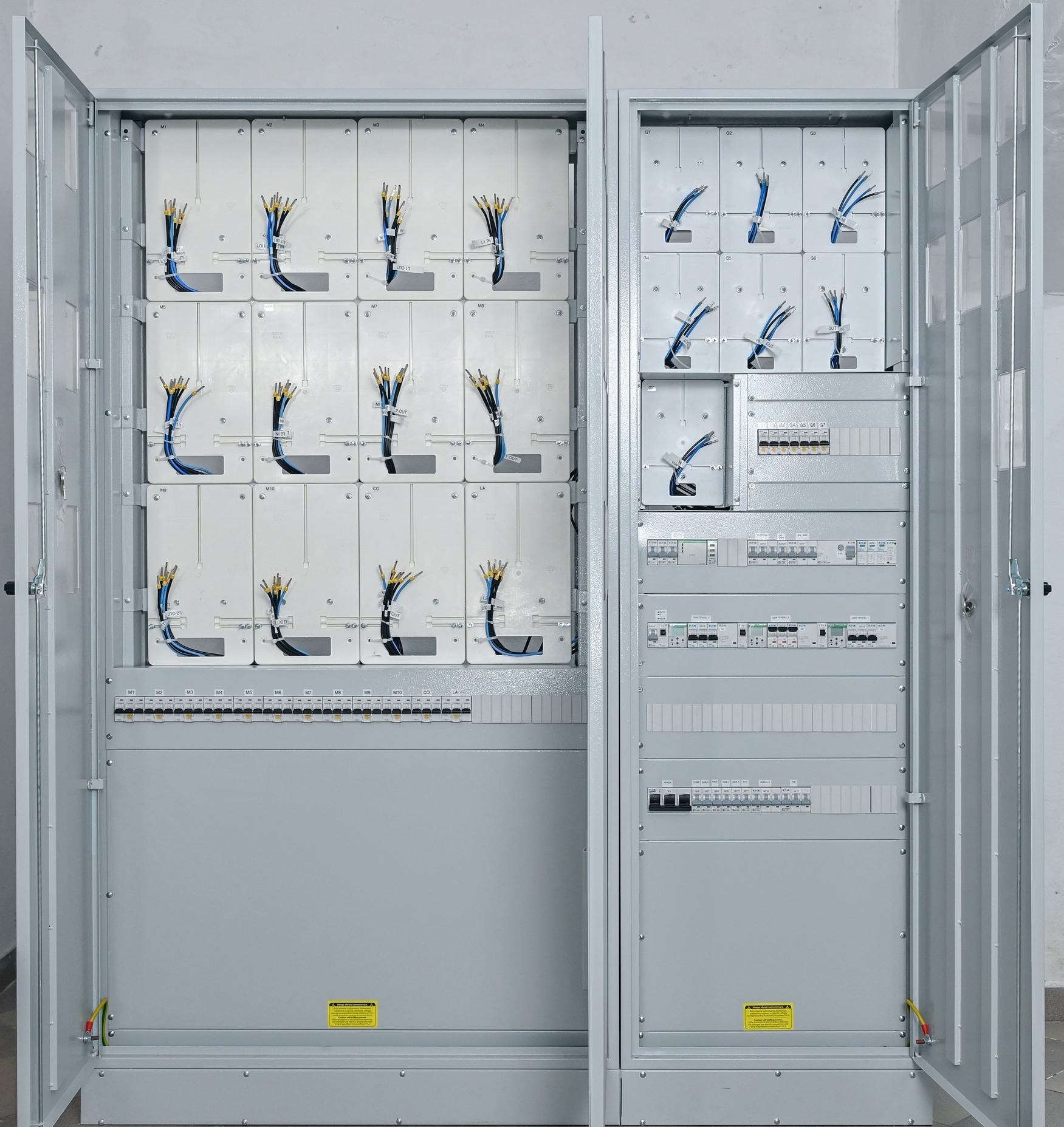 Otwarta szafa sterownicza z panelami elektrycznymi i podłączonymi przewodami, widoczne oznaczenia na panelach i zabezpieczenia elektryczne.