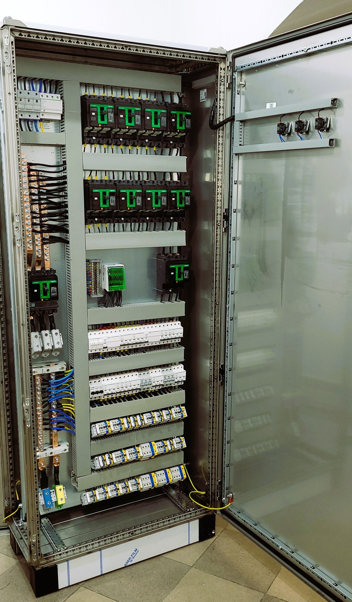 Otwarta szafa sterownicza z widocznymi bezpiecznikami, przekaźnikami i okablowaniem, z żółto-zielonym przewodem uziemiającym w dolnej części.