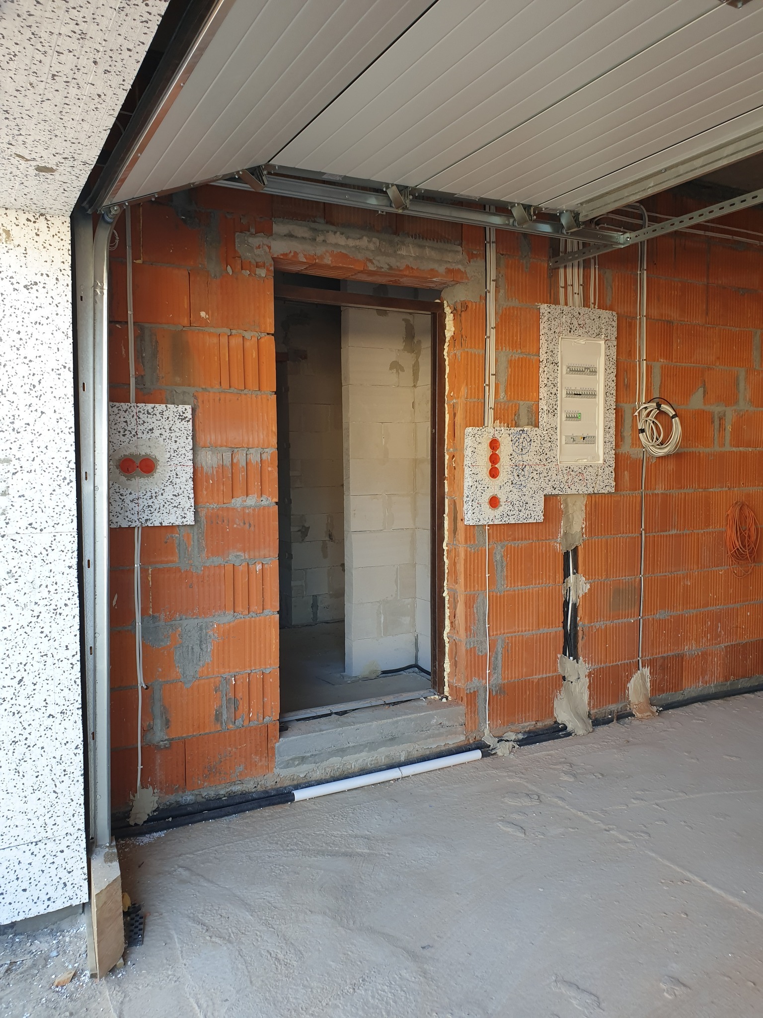 Wnętrze garażu w trakcie budowy z widoczną bramą segmentową, instalacją elektryczną w ścianach z czerwonej cegły oraz skrzynkami bezpiecznikowymi z białymi pokrywami.