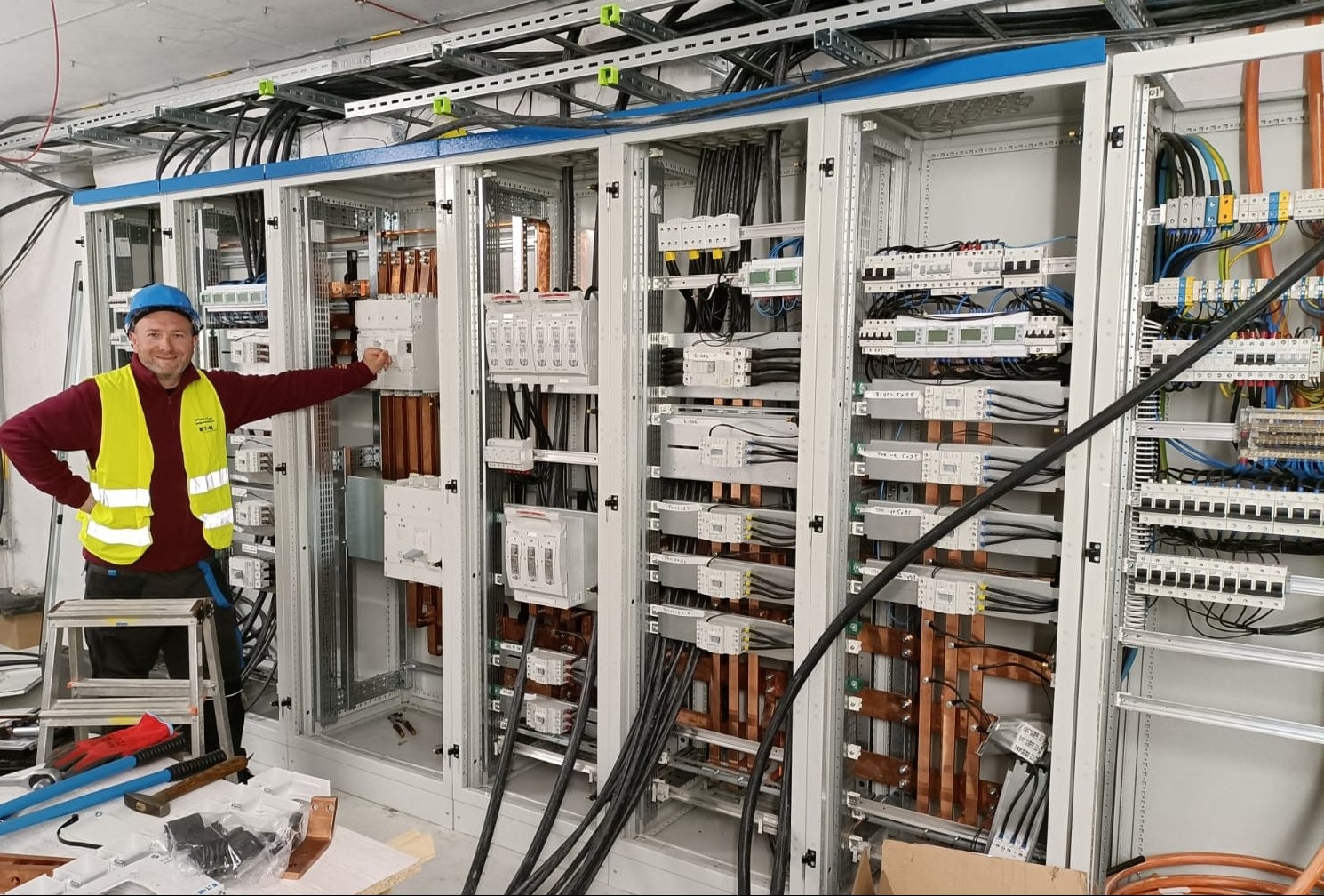 Uśmiechnięty elektryk w kasku i kamizelce odblaskowej prezentuje rozbudowaną szafę rozdzielczą z licznymi przewodami i zabezpieczeniami, widoczną w tle drabina i narzędzia.