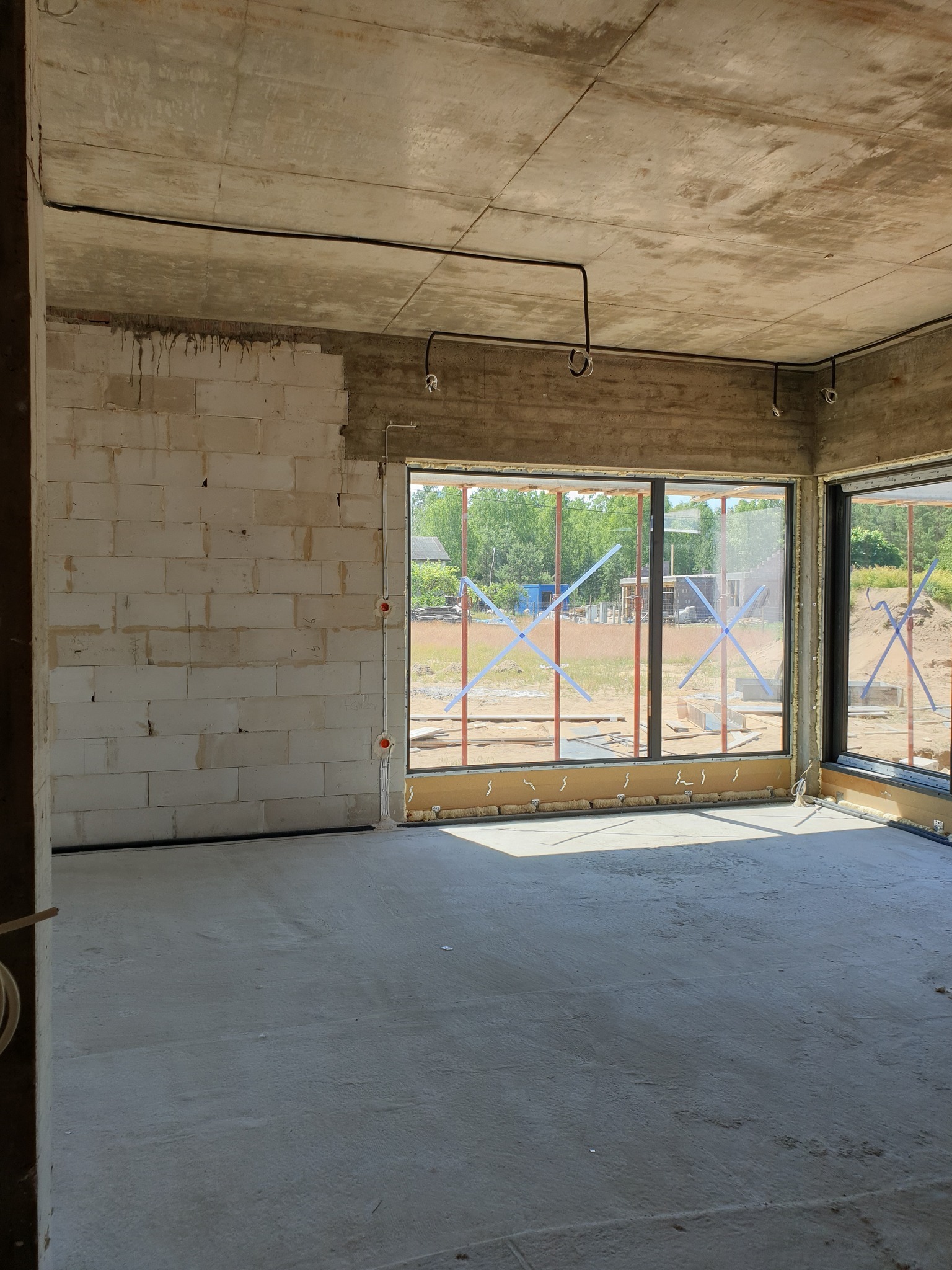 Surowy stan wnętrza budynku z widocznymi przewodami elektrycznymi pod sufitem i puszkami w ścianie z betonu komórkowego, okna zabezpieczone folią.