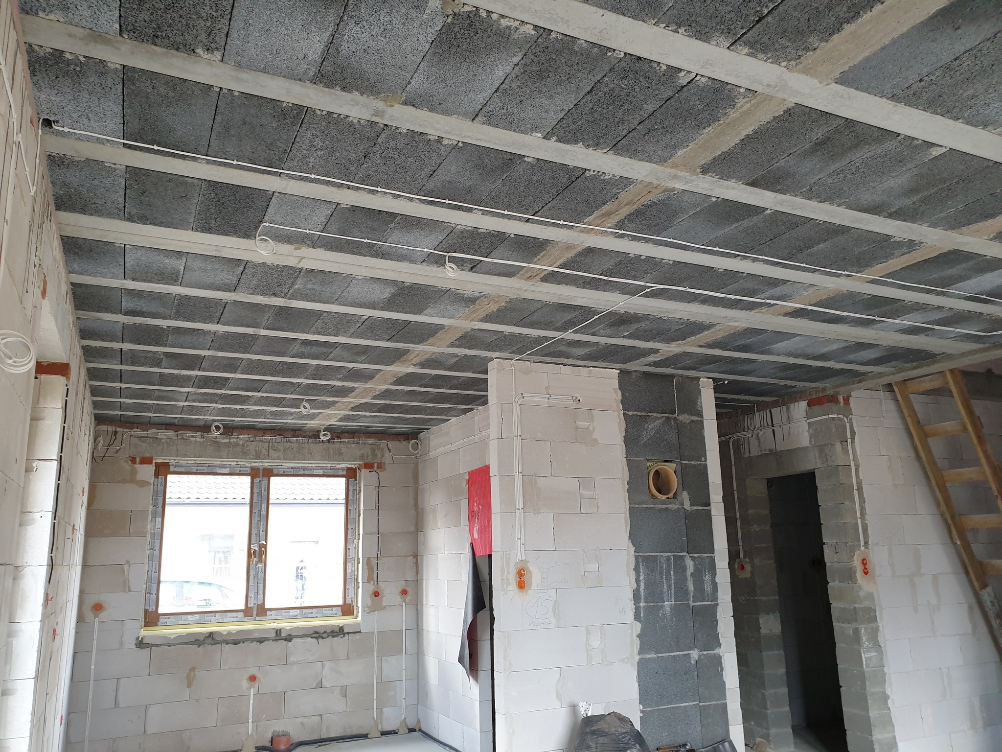 Surowy stan wnętrza budynku w trakcie budowy z widocznymi przewodami elektrycznymi poprowadzonymi pod betonowym stropem i na ścianach z bloczków, okno z widokiem na zewnątrz, surowa ściana...