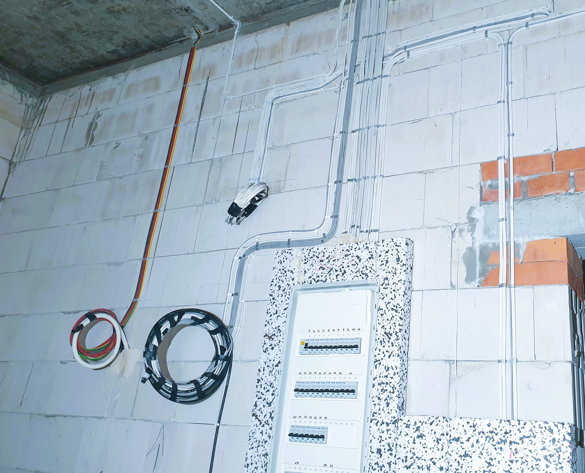 Rozprowadzenie przewodów elektrycznych w peszlach na ścianie z bloczków betonowych, widoczna skrzynka rozdzielcza z bezpiecznikami i zwitki kabli. Prace instalacyjne w surowym stanie.