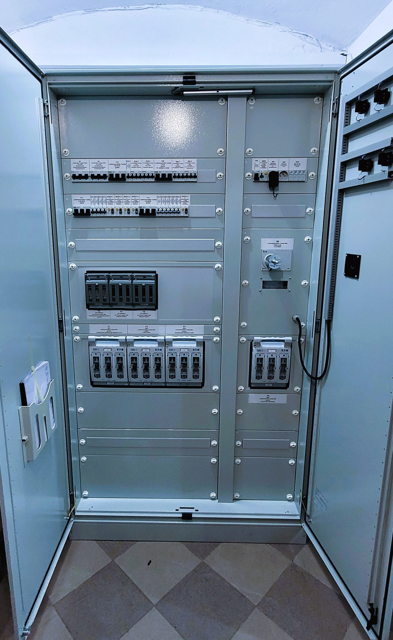 Otwarta szafa elektryczna z widocznymi bezpiecznikami, wyłącznikami i okablowaniem, zamontowana w pomieszczeniu z podłogą w kratkę.