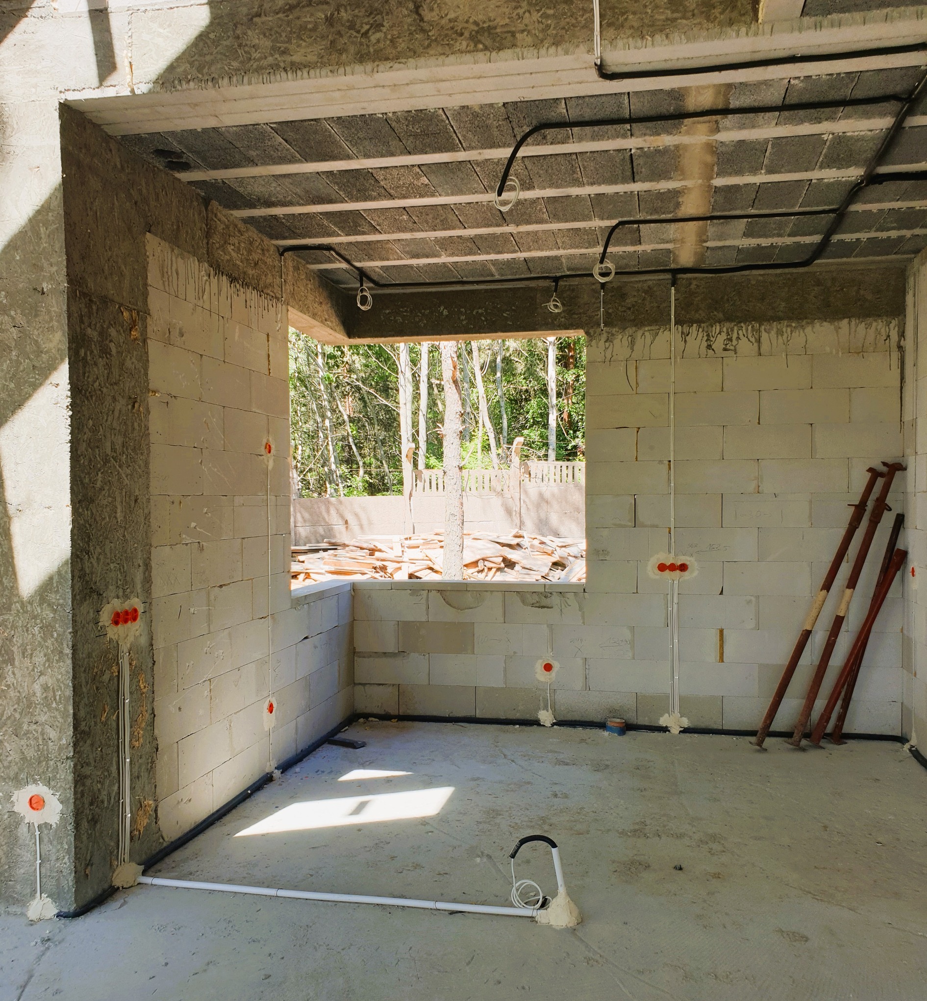 Surowy stan wnętrza budynku w trakcie budowy, widoczne instalacje elektryczne w ścianach z betonu komórkowego oraz przewody pod sufitem, przez okno widok na drzewa.