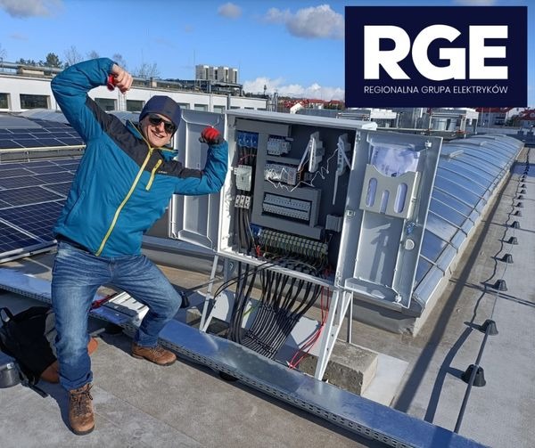 Elektryk w niebieskiej kurtce i czapce, z uniesionymi rękami w geście triumfu, stoi na dachu obok otwartej skrzynki elektrycznej z widocznymi przewodami i elementami sterującymi; w tle panele...