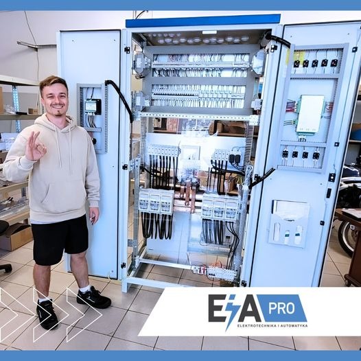 Młody mężczyzna w bluzie i spodenkach prezentuje otwartą szafę sterowniczą z okablowaniem i elementami automatyki, gestykulując dłonią w znak OK.