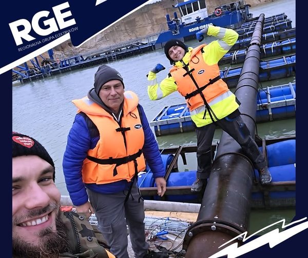 Trzech mężczyzn w kamizelkach ratunkowych pozuje na tle rurociągu pływającego po zbiorniku wodnym, z widoczną pogłębiarką i brzegiem w tle, logo firmy w rogu.