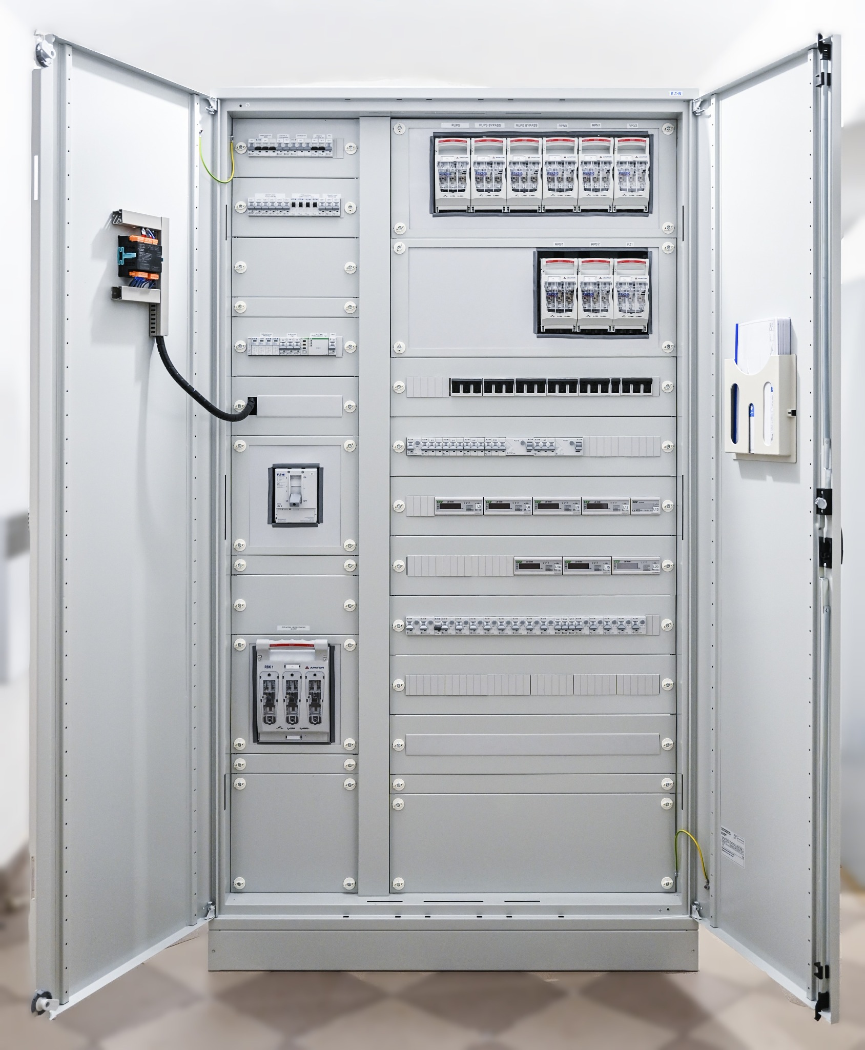 Otwarta szafa elektryczna z widocznymi zabezpieczeniami, wyłącznikami i listwami zaciskowymi, zamontowana w jasnym pomieszczeniu.