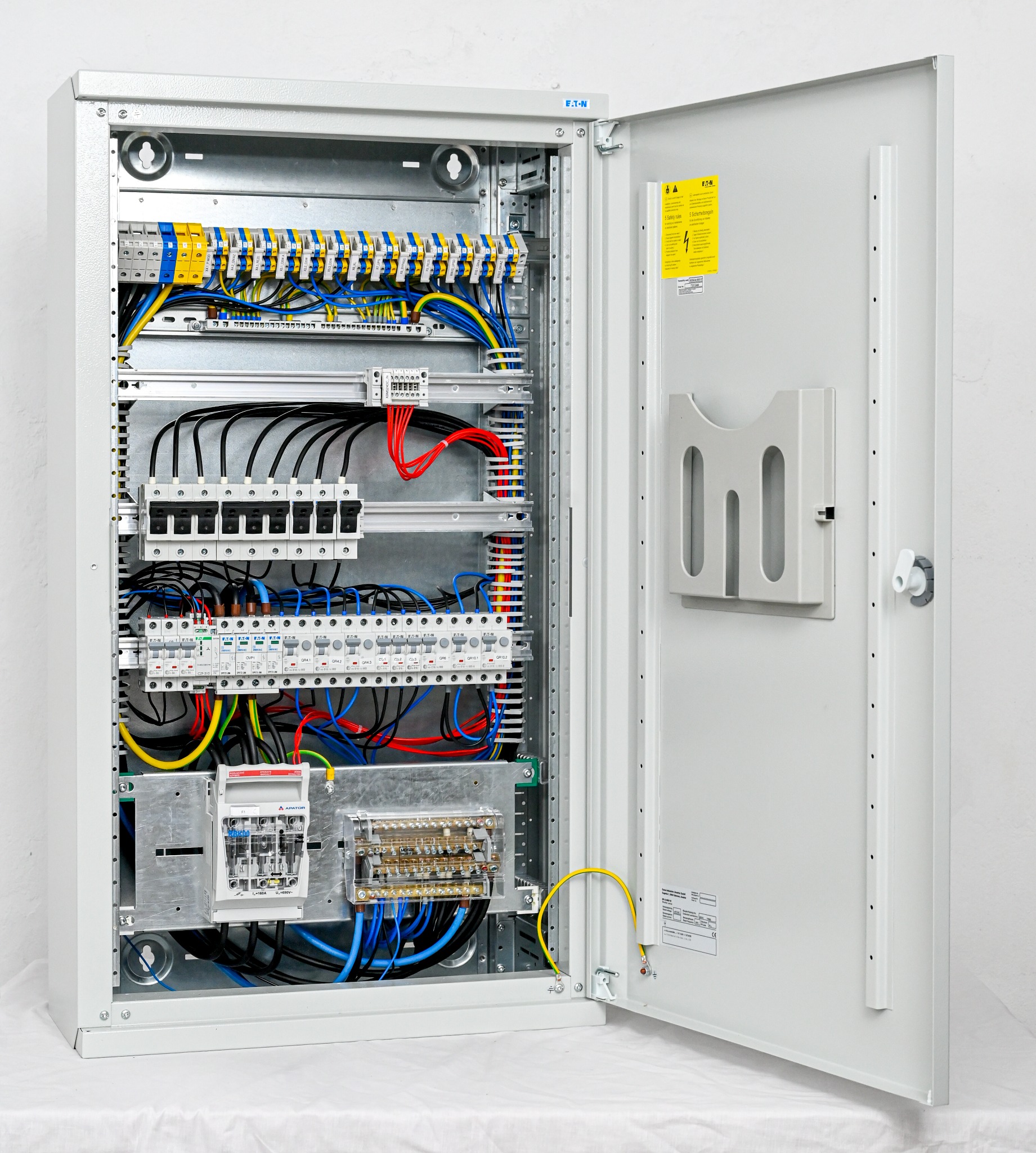 Otwarta, metalowa szafa elektryczna z widocznymi bezpiecznikami, przewodami w różnych kolorach oraz elementami montażowymi, zamontowana na białym tle.