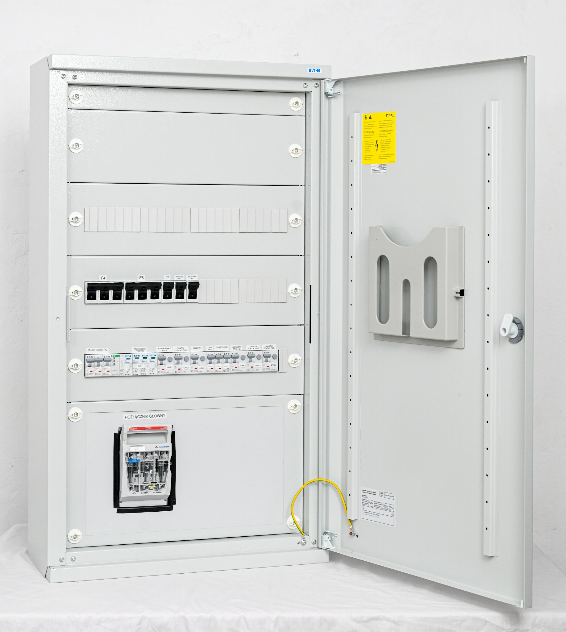 Otwarta szafa elektryczna z widocznymi bezpiecznikami, wyłącznikami różnicowoprądowymi i żółto-zielonym przewodem uziemiającym, zamontowana na białym tle.