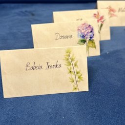 Joanna Pytka-Piecychna - Ręcznie robione winietki z imionami gości i motywami kwiatowymi, ustawione rzędami na niebieskim tle, w delikatnym oświetleniu.