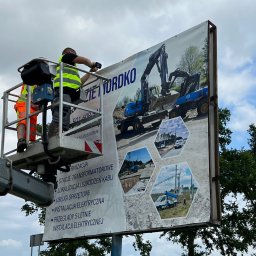 Joanna Pytka-Piecychna - Montaż baneru reklamowego z grafiką koparek i usług elektrycznych przez dwóch pracowników na podnośniku koszowym na tle pochmurnego nieba i drzew.