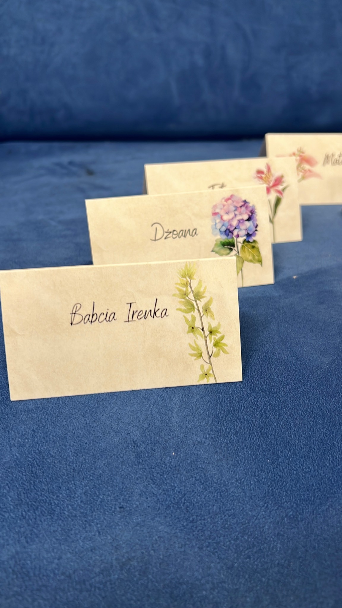 Ręcznie robione winietki z imionami gości i motywami kwiatowymi, ustawione rzędami na niebieskim tle, w delikatnym oświetleniu.