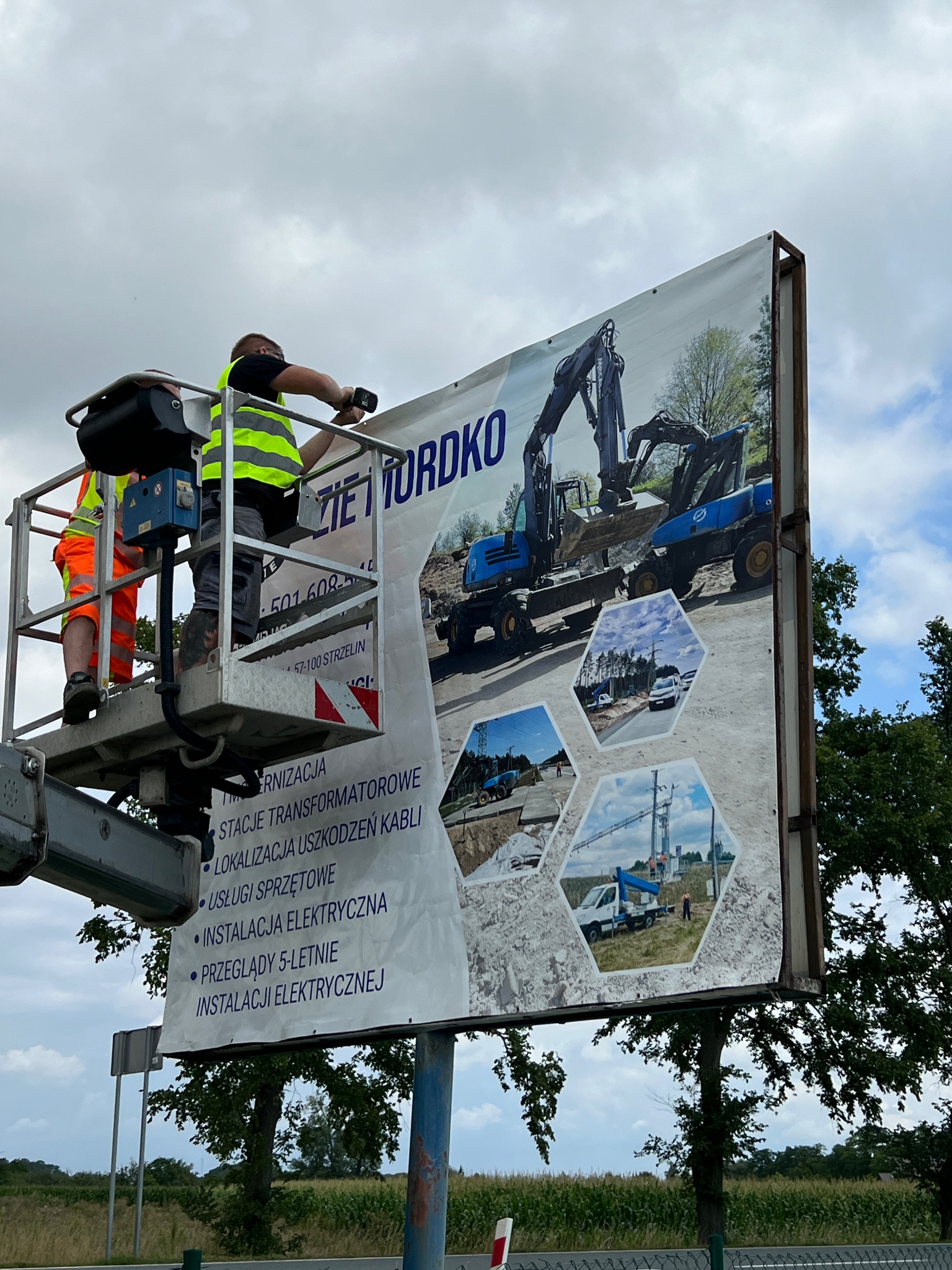 Montaż baneru reklamowego z grafiką koparek i usług elektrycznych przez dwóch pracowników na podnośniku koszowym na tle pochmurnego nieba i drzew.