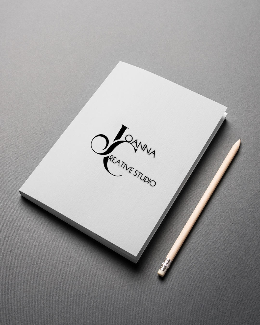 Elegancki notes z logo 'Joanna Creative Studio' leżący na szarym tle obok ołówka. Minimalistyczny design, czcionka szeryfowa, stonowana kolorystyka.
