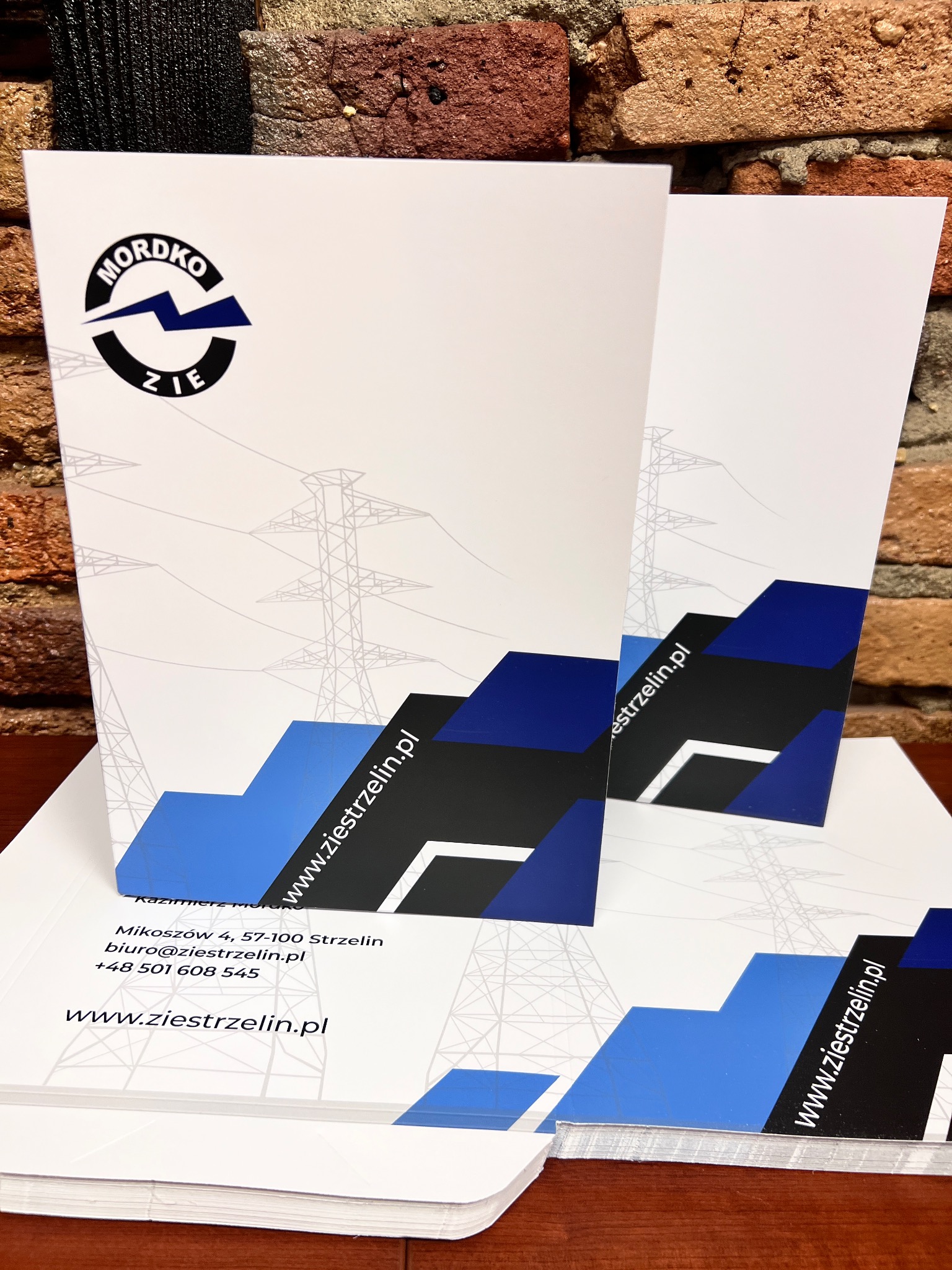 Stos firmowych papierów firmowych z logo i danymi kontaktowymi ZIE Mordko, umieszczonych na tle ceglanej ściany. Widoczne motywy słupów wysokiego napięcia.
