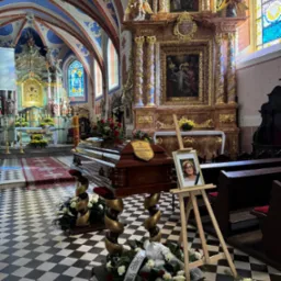 Wnętrze kościoła z drewnianą trumną na ozdobnych stojakach, otoczoną wieńcami z białych róż, obok portret z czarną wstążką na sztaludze.