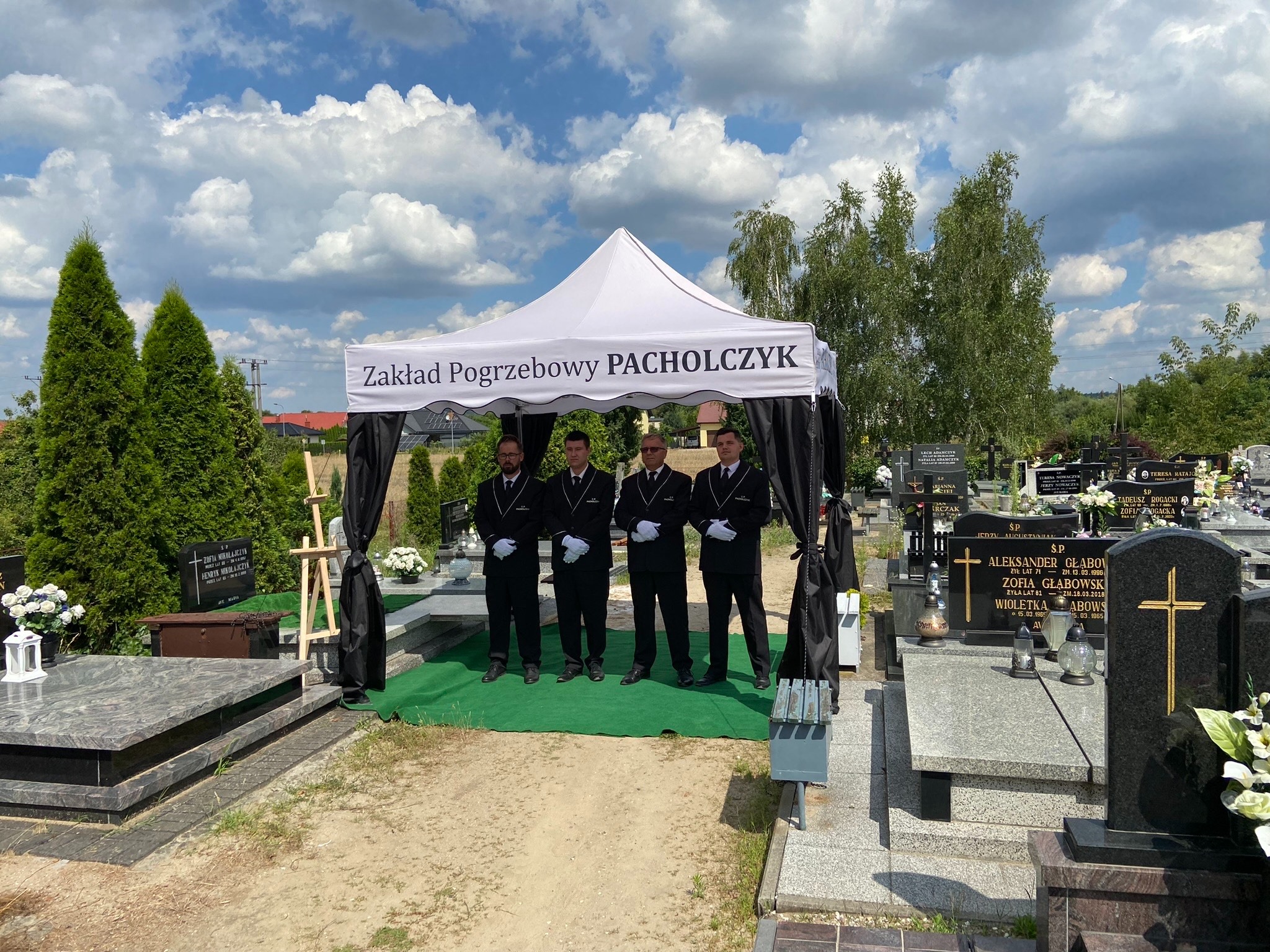 Czterej mężczyźni w garniturach i białych rękawiczkach stoją pod białym namiotem z napisem 'Zakład Pogrzebowy PACHOLCZYK' na tle cmentarza z nagrobkami.