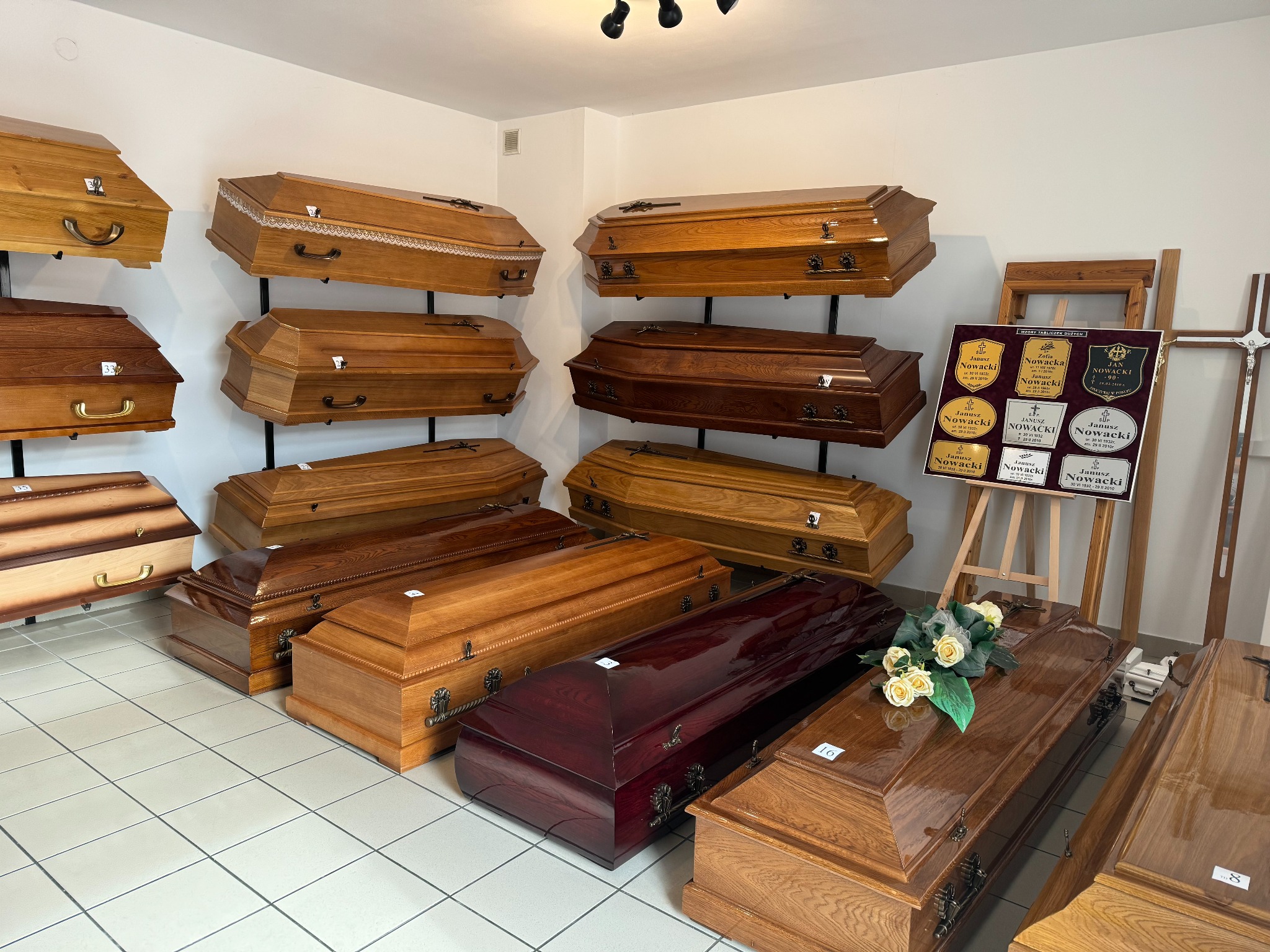 Ekspozycja trumien w zakładzie pogrzebowym: drewniane trumny w różnych odcieniach brązu, niektóre z dekoracyjnymi elementami metalowymi i krzyżami, oraz tabliczki nagrobne na sztalugach w tle.