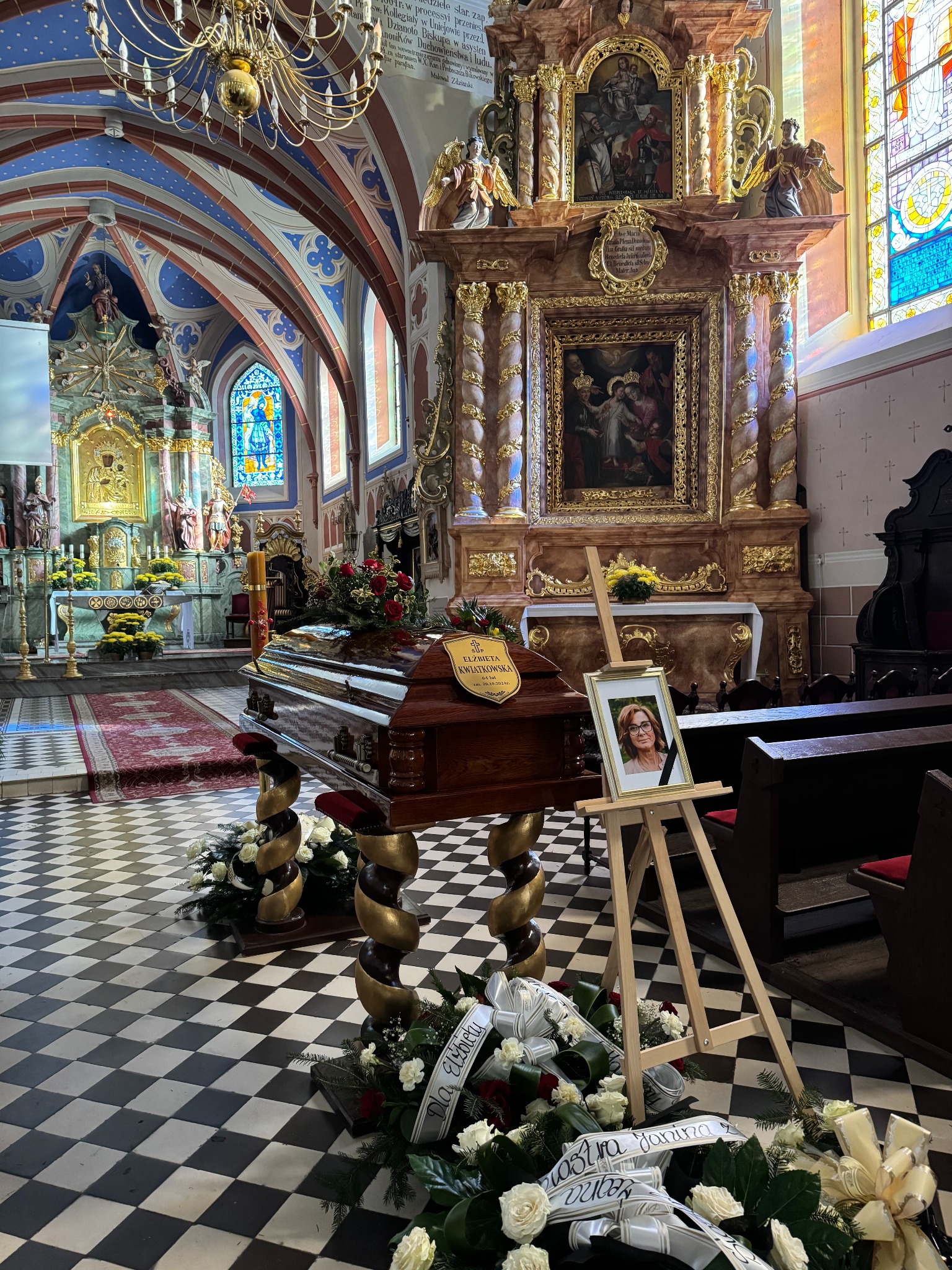 Wnętrze kościoła z drewnianą trumną na ozdobnych stojakach, otoczoną wieńcami z białych róż, obok portret z czarną wstążką na sztaludze.