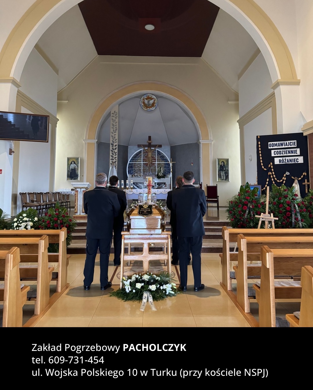 Wnętrze kościoła z trumną na podeście, otoczoną wieńcami pogrzebowymi i kwiatami, widok z perspektywy nawy głównej, w tle krzyż i ołtarz.
