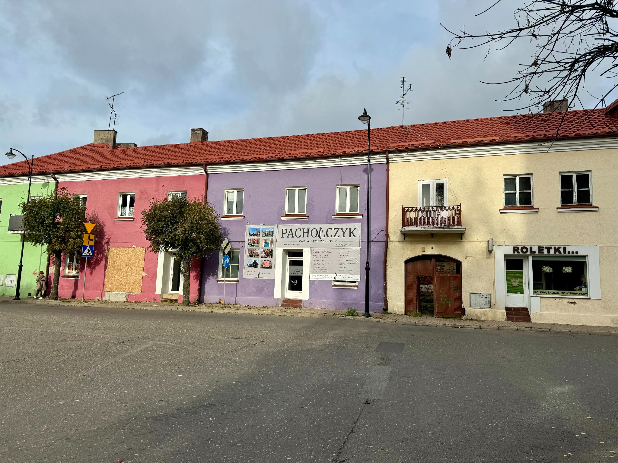 Kolorowe kamienice w rzędzie, w tym fioletowa z szyldem zakładu pogrzebowego 'PACHOLCZYK', obok budynek z reklamą roletek; ulica przed budynkami.