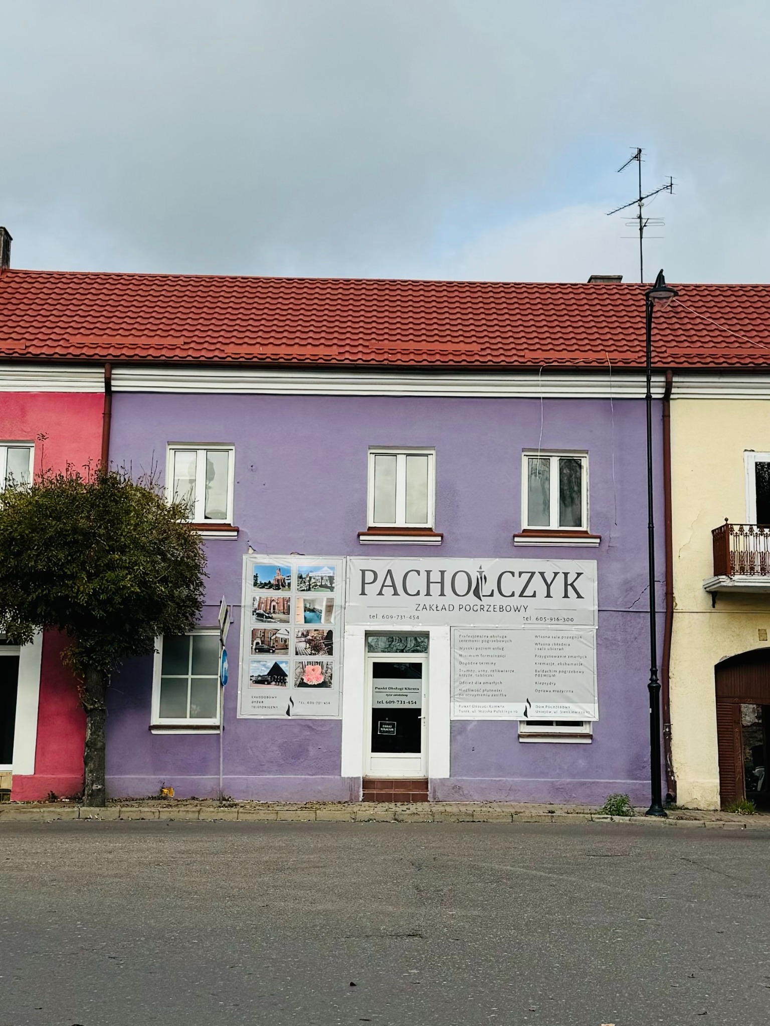 Fioletowa fasada Zakładu Pogrzebowego Pacholczyk z reklamami usług pogrzebowych w oknach, czerwony dach, pochmurne niebo.