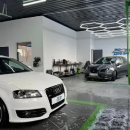 Dwa samochody w myjni, białe Audi z przodu i szary Seat Ibiza z tyłu, mokra podłoga z zielonymi liniami, oświetlenie sufitowe w kształcie plastra miodu, widoczne butelki z detergentami na półkach.