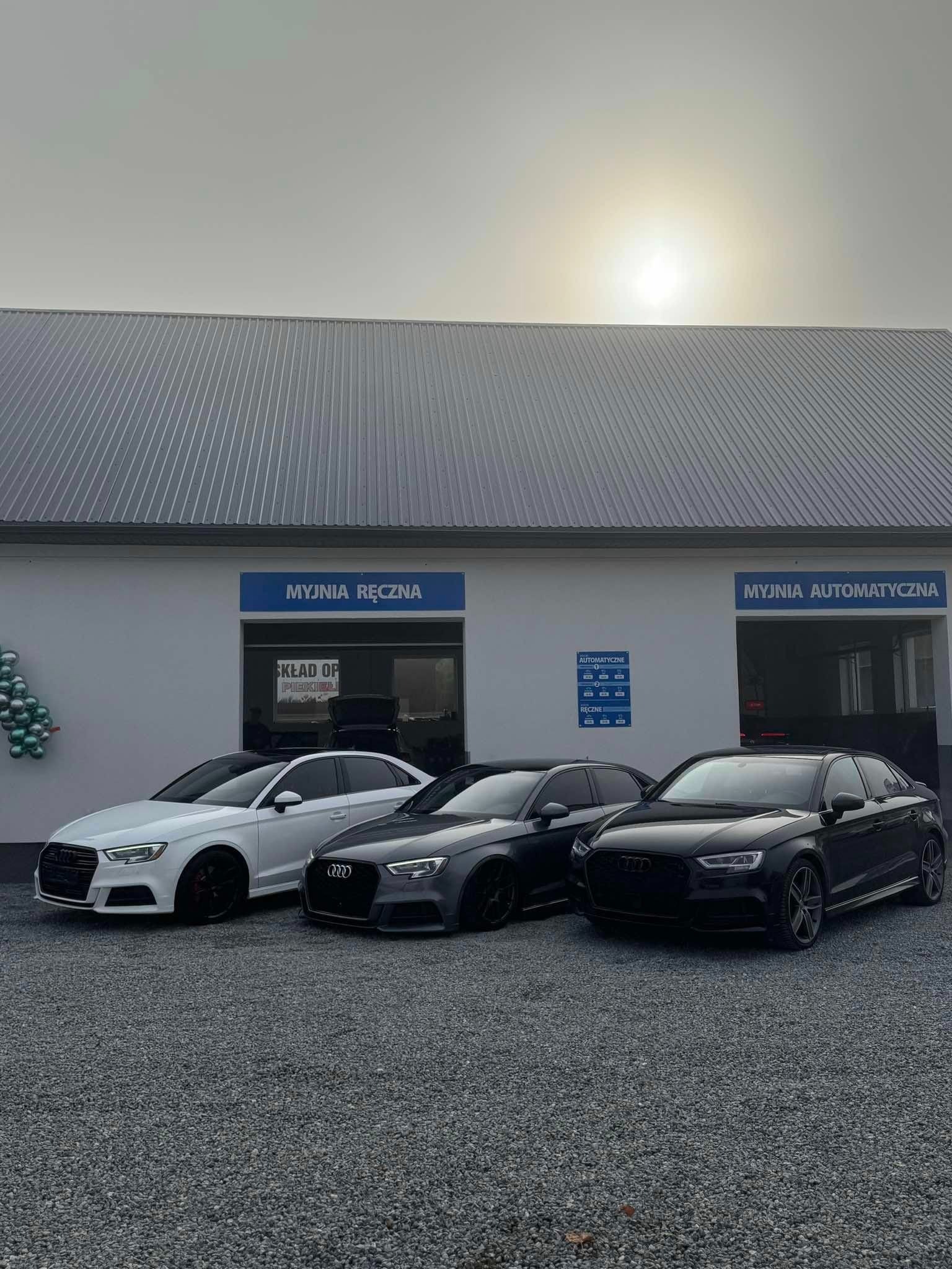 Trzy Audi A3 w kolorach białym, szarym i czarnym zaparkowane przed budynkiem myjni z napisem 'Myjnia Ręczna' i 'Myjnia Automatyczna' w pochmurny dzień.