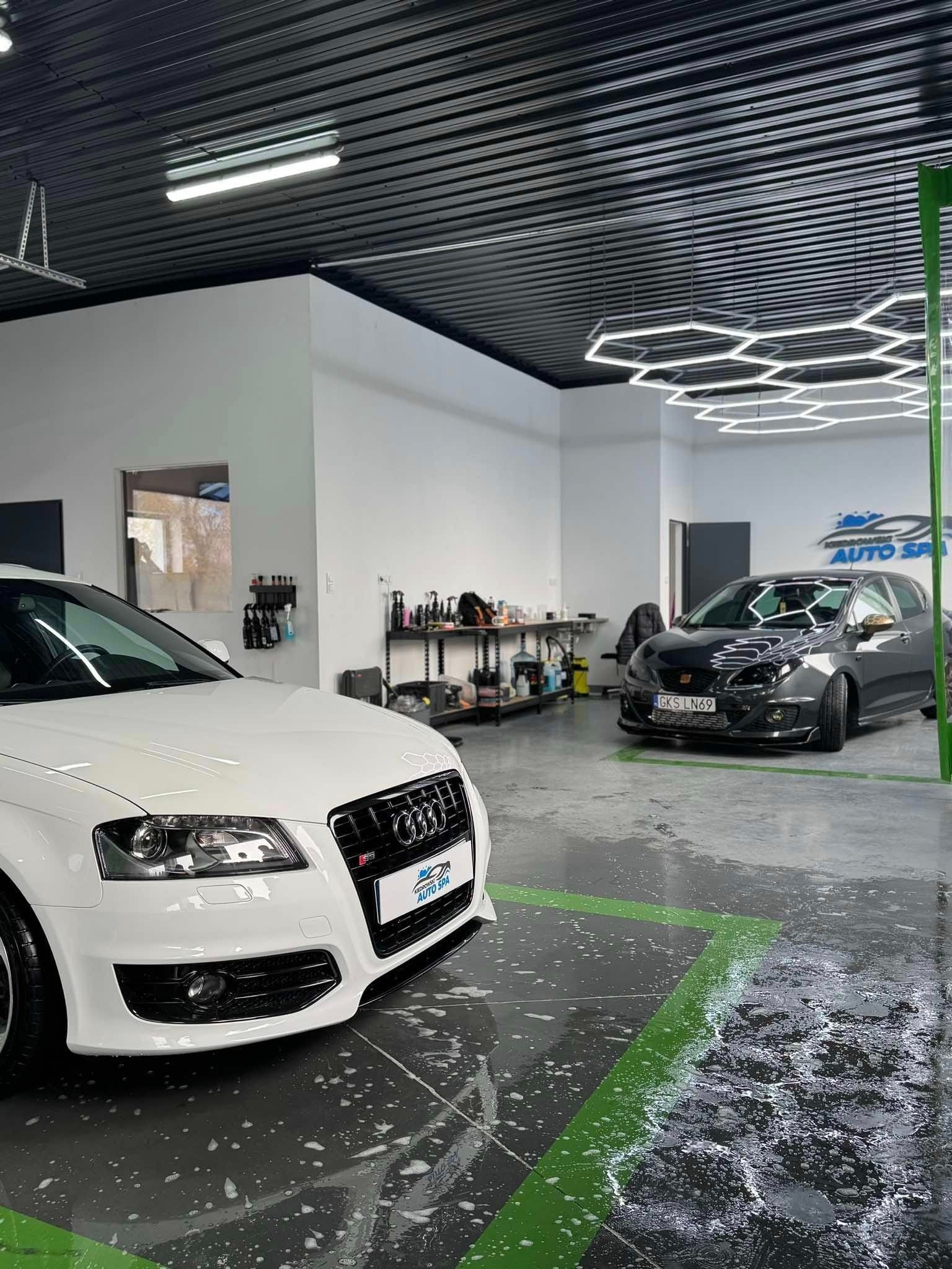 Dwa samochody w myjni, białe Audi z przodu i szary Seat Ibiza z tyłu, mokra podłoga z zielonymi liniami, oświetlenie sufitowe w kształcie plastra miodu, widoczne butelki z detergentami na półkach.