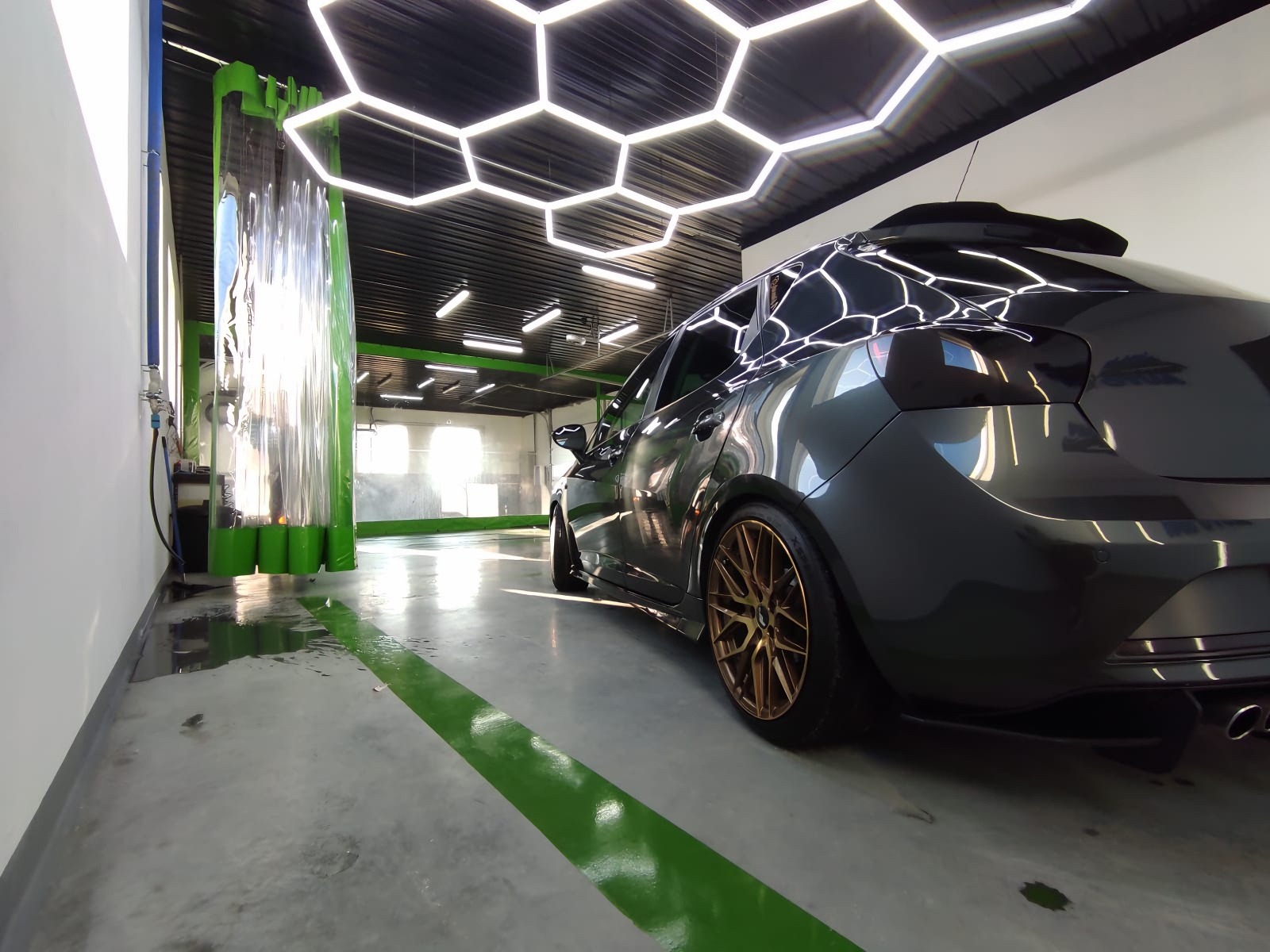 Ciemny samochód po umyciu, z widocznymi kroplami wody na lakierze, stoi w nowoczesnej myjni z oświetleniem w kształcie plastra miodu na suficie i zielonymi akcentami.