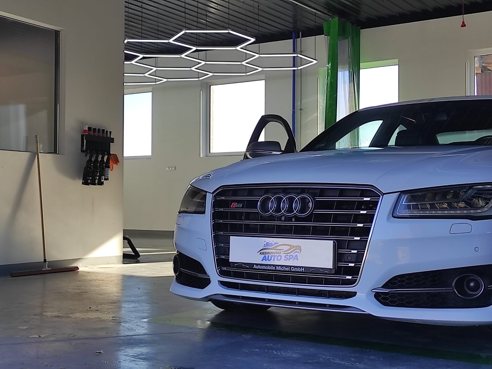 Białe Audi S8 w jasnym wnętrzu myjni samochodowej, z otwartymi drzwiami i widocznym logo firmy detailingowej na grillu.