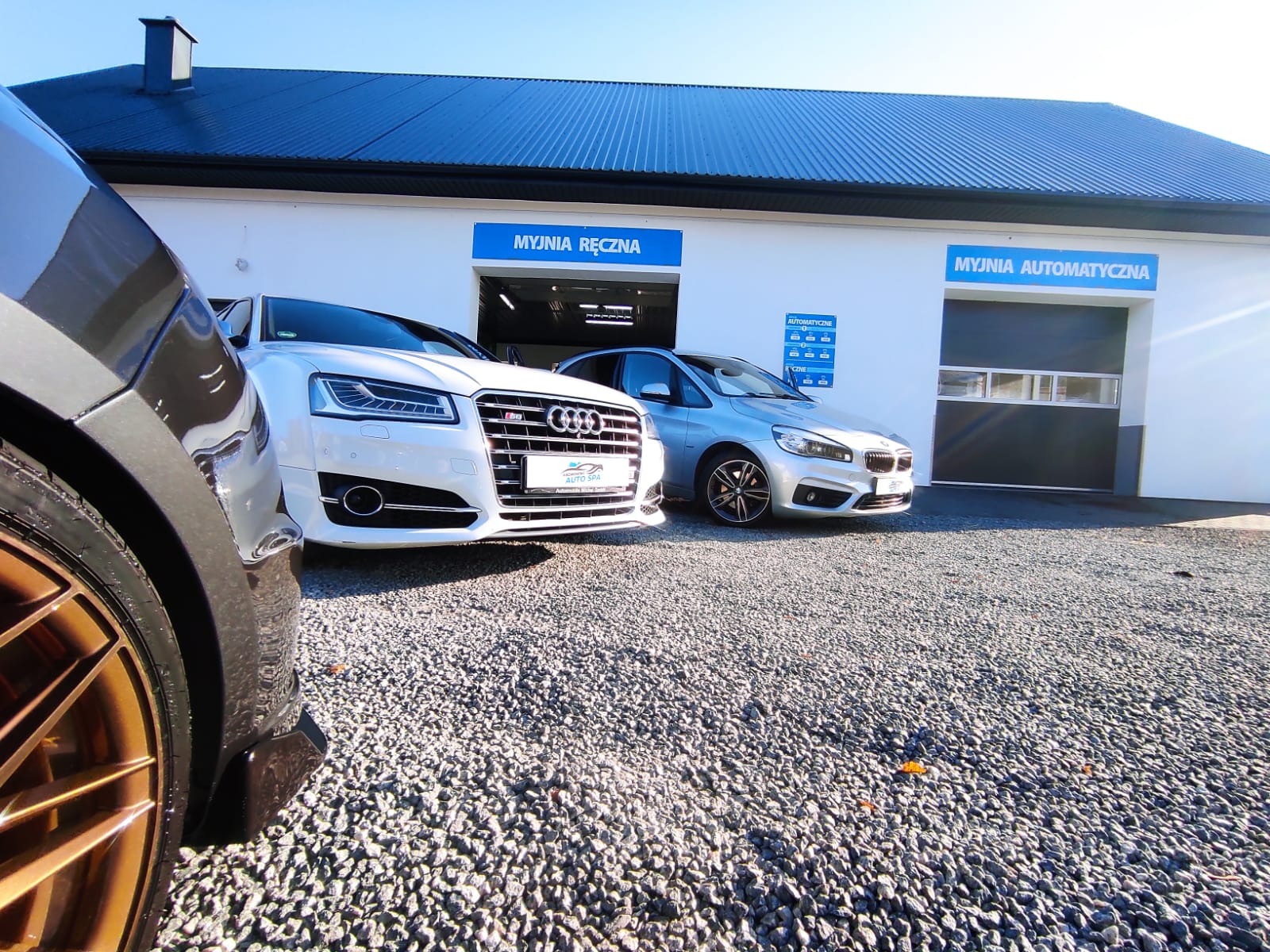Dwa samochody Audi i BMW zaparkowane przed budynkiem myjni z napisem 'Myjnia Ręczna' i 'Myjnia Automatyczna', z widocznym fragmentem trzeciego auta z przodu po lewej stronie.