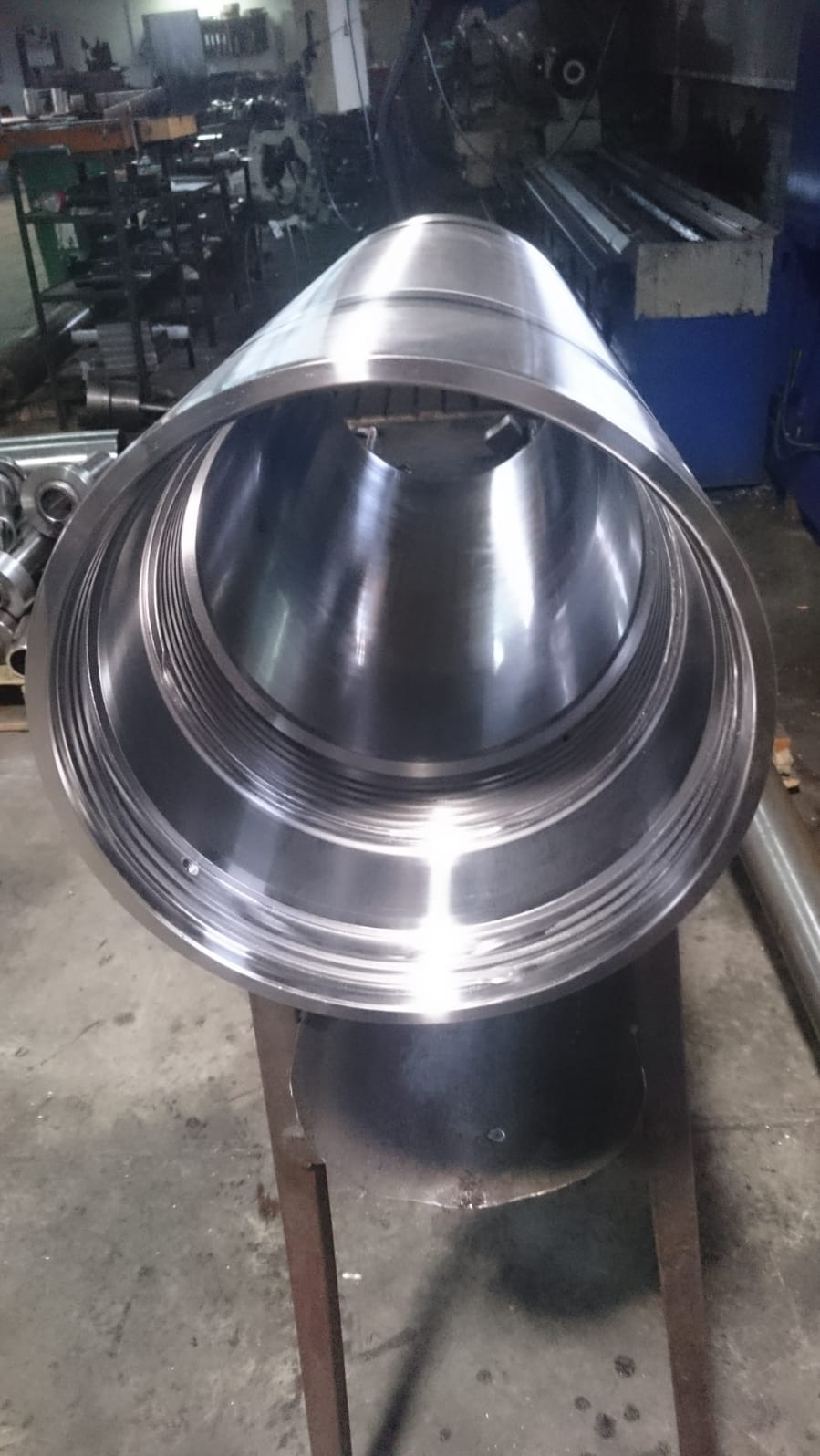 Polerowany, cylindryczny element metalowy z wewnętrznymi gwintami, umieszczony na metalowym stojaku w warsztacie.