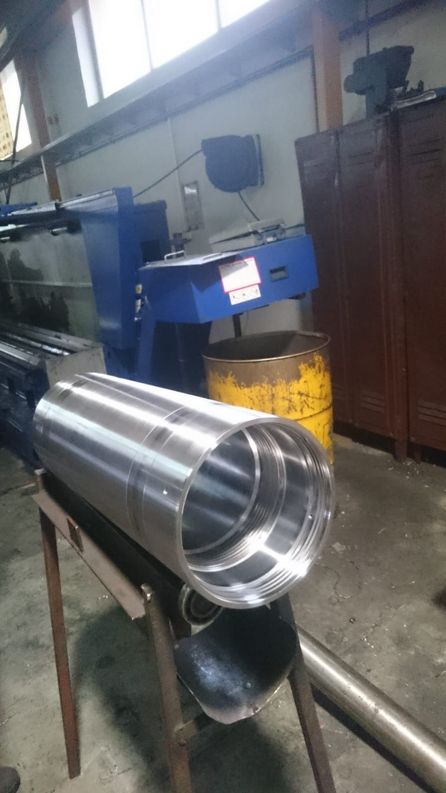 Polerowany metalowy cylinder z gwintem wewnętrznym, leżący na metalowym stojaku, z maszyną w tle i żółtą beczką obok.