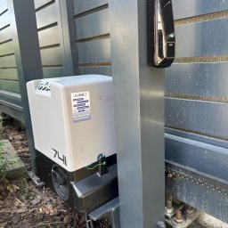 Jarmet - Bramy garażowe Wiśniowski - Szary automat bramy przesuwnej Jarmet 741 z fotokomórką FAAC, zamontowany przy ogrodzeniu z poziomych paneli. Widoczne elementy montażowe i podłoże.