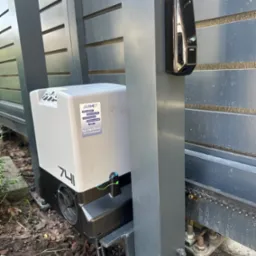 Szary automat bramy przesuwnej Jarmet 741 z fotokomórką FAAC, zamontowany przy ogrodzeniu z poziomych paneli. Widoczne elementy montażowe i podłoże.