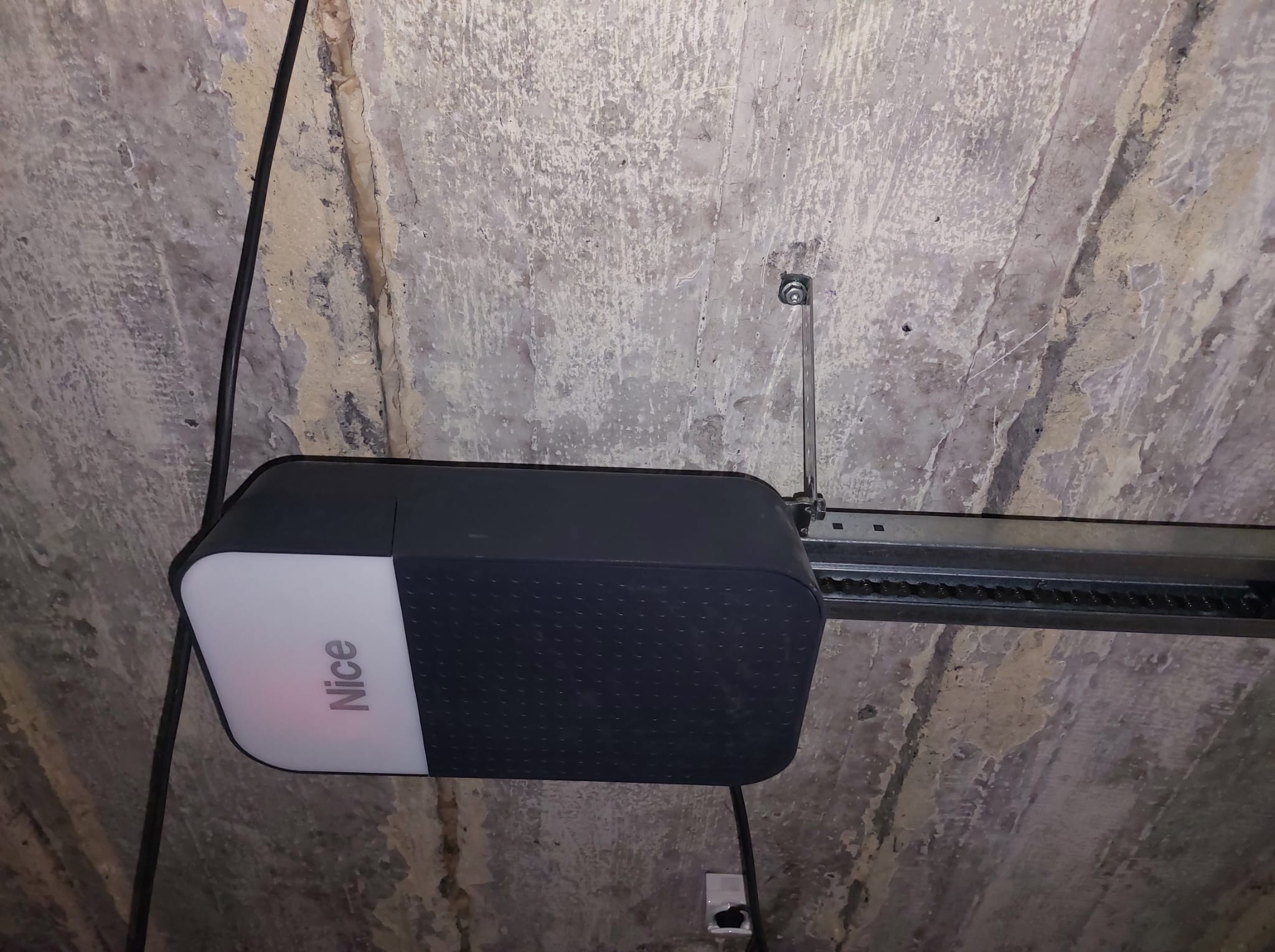 Zamontowany napęd do bramy garażowej marki Nice, widoczny mechanizm łańcuchowy i przewody, tło stanowi surowy betonowy sufit garażu.