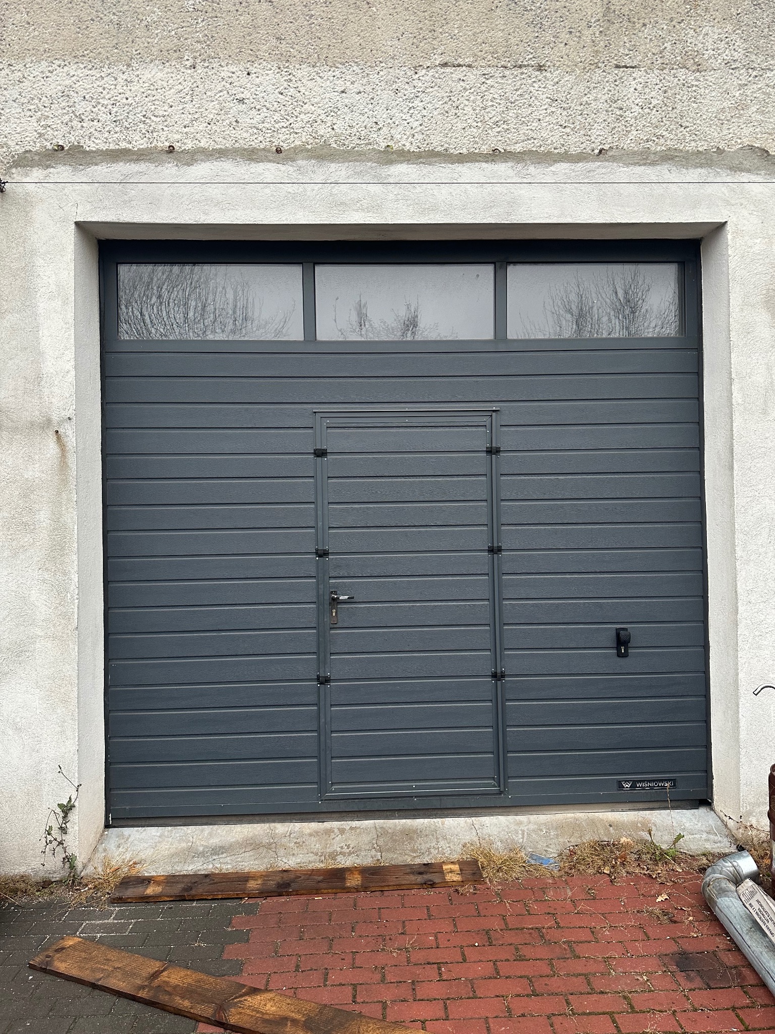 Antracytowa brama garażowa segmentowa Wiśniowski z niskim przetłoczeniem woodgrain, wyposażona w drzwi serwisowe, okucia w kolorze bramy i trzy panele przeszklone u góry, zamontowana w starej...