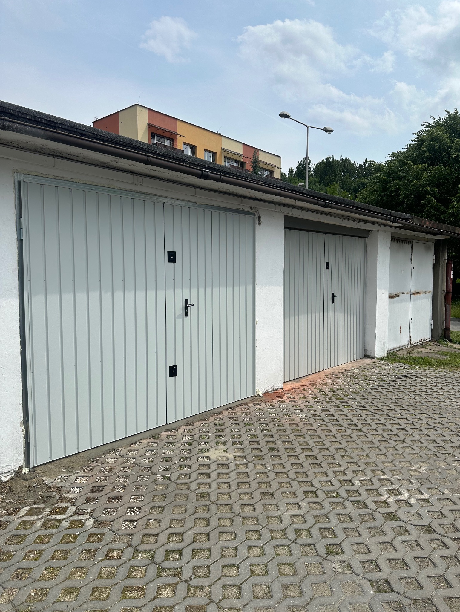 Dwie garażowe bramy segmentowe, jedna w kolorze szarym z czarnymi uchwytami, druga biała, z widoczną kostką brukową i budynkiem w tle.