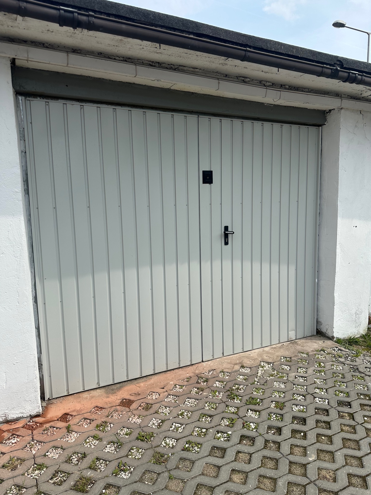 Szara, dwuskrzydłowa brama garażowa z czarną klamką, zamontowana w białym, murowanym garażu. Kostka brukowa przed bramą.