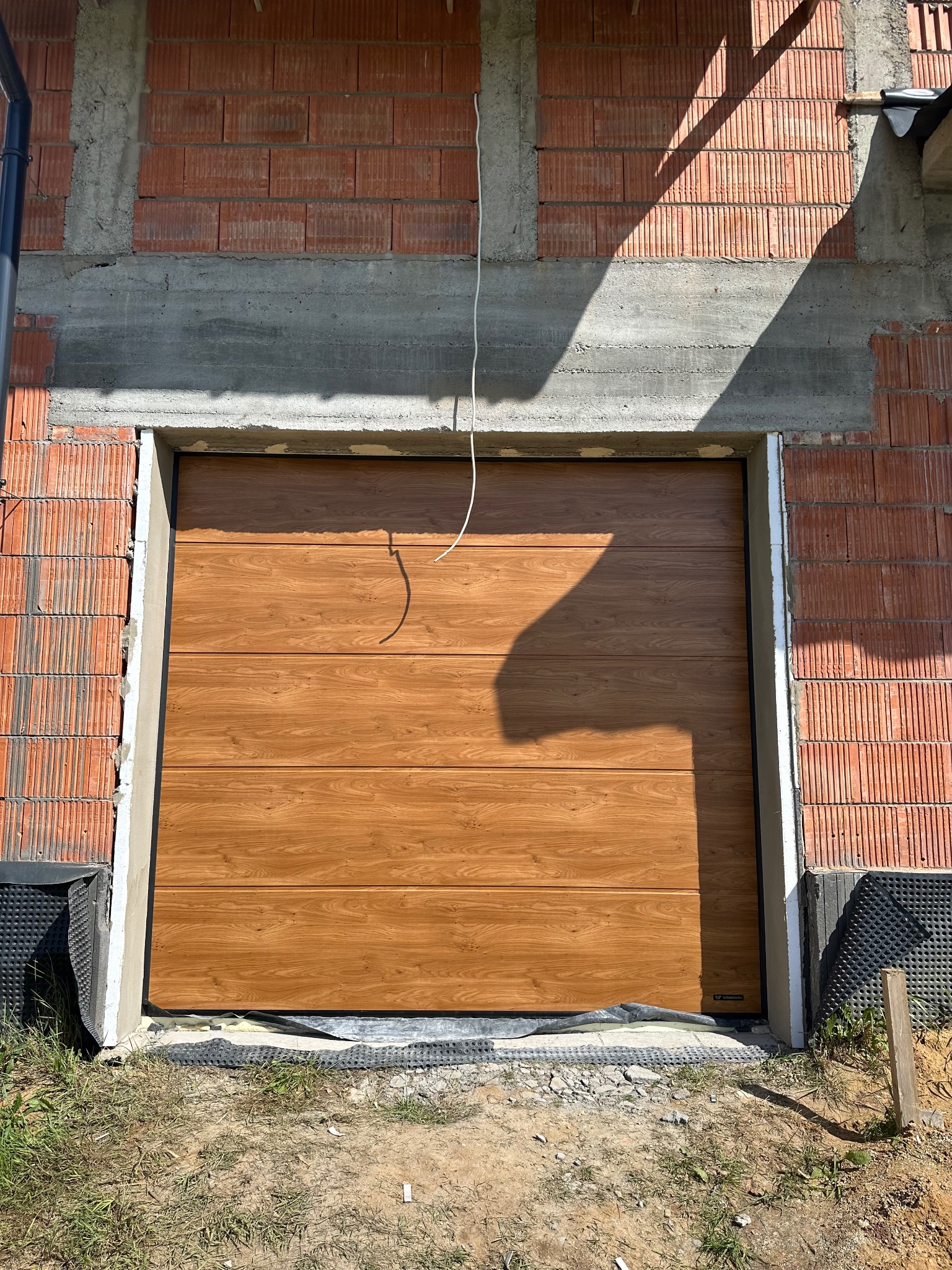 Zamknięta, segmentowa brama garażowa w odcieniach brązu, zamontowana w surowym otworze budynku z czerwonej cegły. Widoczne oświetlenie słoneczne i fragmenty instalacji.