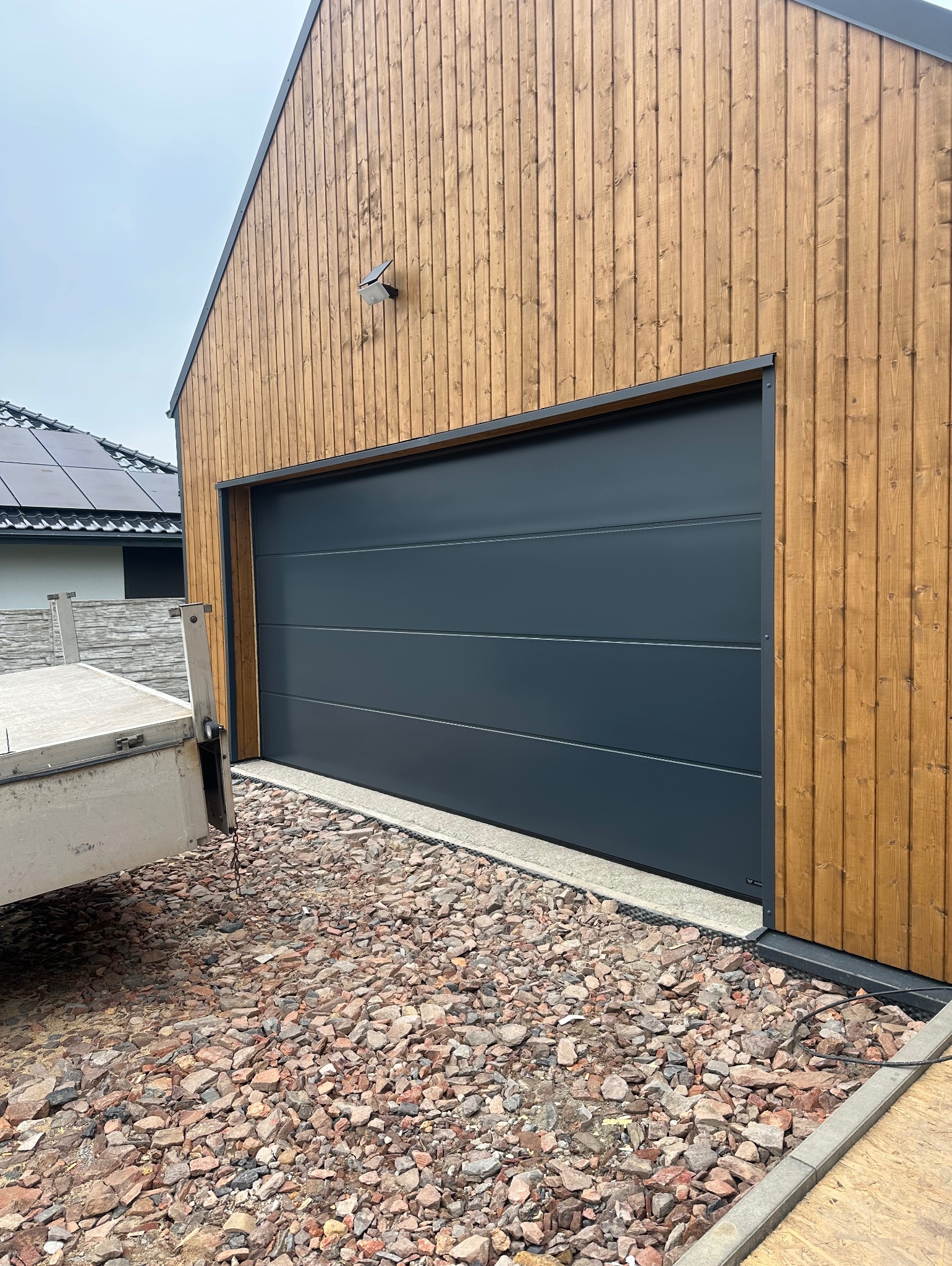 Zamknięta, segmentowa brama garażowa w kolorze antracytowym, zamontowana w budynku z drewnianą elewacją. Widoczne kamienie ozdobne przed bramą.