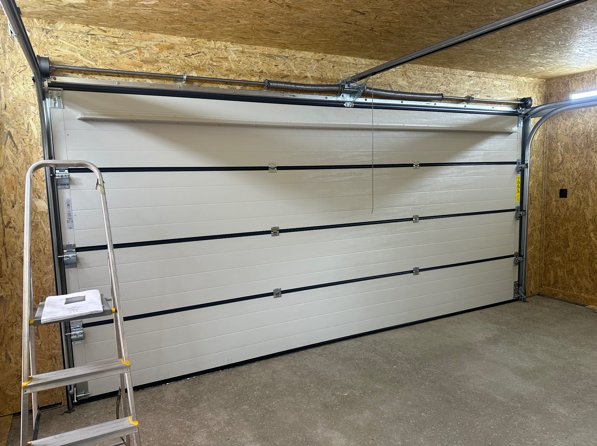 Zamknięta, segmentowa brama garażowa w kolorze białym, z widocznym mechanizmem sprężynowym i drabinką. Ściany garażu wyłożone płytami OSB.
