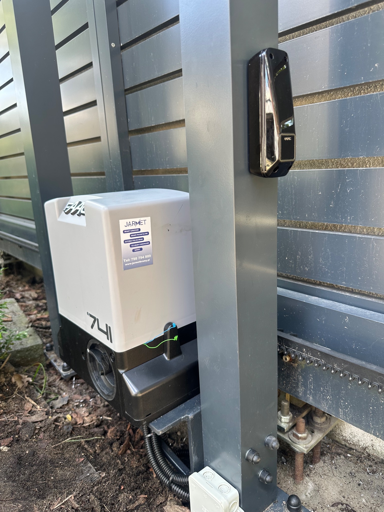 Szary automat bramy przesuwnej Jarmet 741 z fotokomórką FAAC, zamontowany przy ogrodzeniu z poziomych paneli. Widoczne elementy montażowe i podłoże.