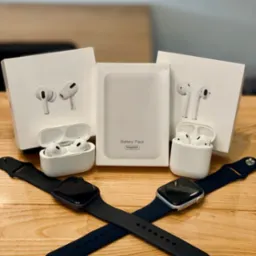 Dwa skrzyżowane smartwatche leżące na drewnianym blacie, otoczone pudełkami bezprzewodowych słuchawek i baterią MagSafe; prezentacja produktów Apple.